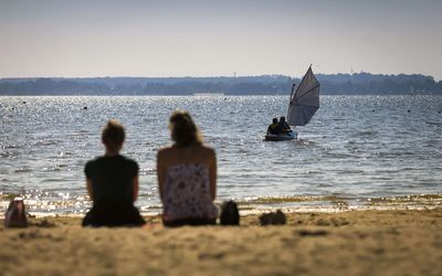 Boot und Strand Zwei Frauen geniessen ihre Freizeit am Badestrand in Mardorf und schauen zwei Segeln in einem kleinen Boot auf dem Wasser zu