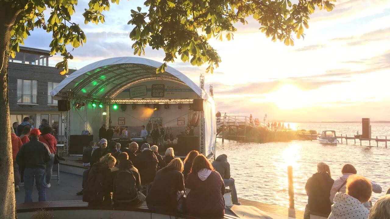 Auf der Seebühne im Hafen von Steinhude steht eine Band im Sonnenuntergang, davor viele Zuschauer