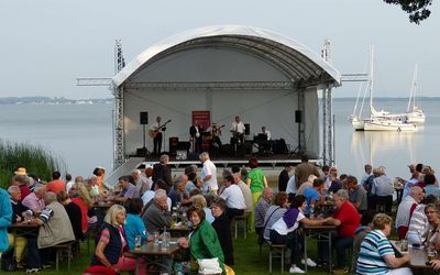 Seebühne in Mardorf am Ufer der Promenade Zahlreiches Publikum hat sich an einem lauen Sommerabend in Mardorf vor der Seebühne versammelt und lauscht dem Konzert. Im Hintergrund dümpeln Segelboote auf dem spiegelnden Wasser.