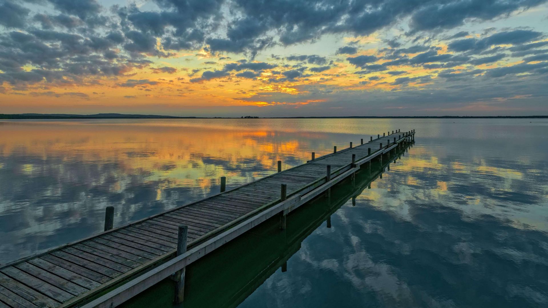 Die Sonne geht orangefarbend am wolkigen Abendhimmel unter. Im Vordergrund ist ein langer Steg .
