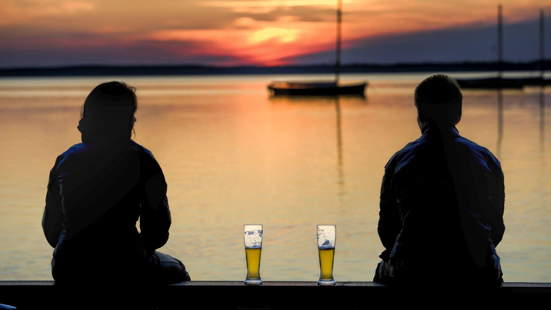 Ein Paar sitzt mit zwei Weizenbiergläsern zwischen sich an der Uferkante am Steinhuder Meer und schaut in den Sonnenuntergang
